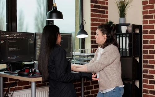 An image showing two women huddled before a dual monitor computer while coding.