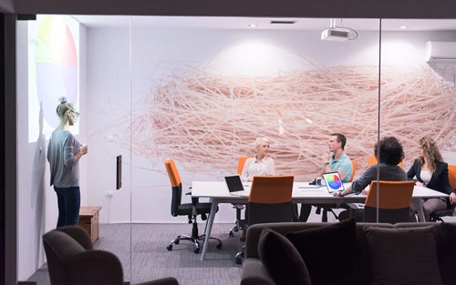 A photo showing a standing woman discussing in front of her seated coworkers inside a glass walled meeting room.
